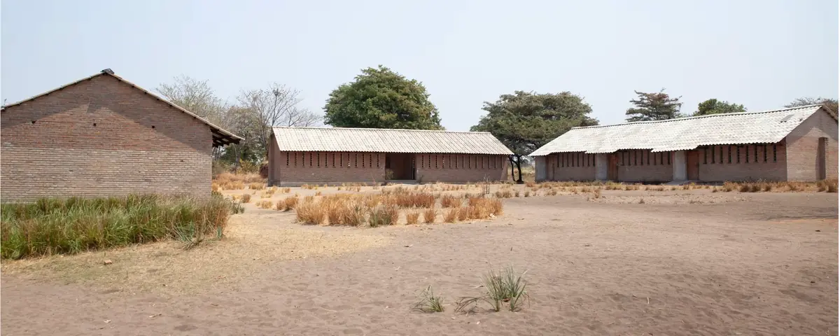Three brick classroom blocks with pale roofs around a sandy school yard, with trees behind