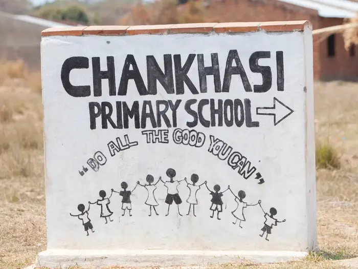 The painted school sign: Chankhasi Primary School, with the motto Do all the good you can above a drawing of children holding hands